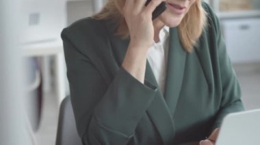 Orta yaşlı, beyaz ceketli bir iş kadınının, ofiste otururken ve cep telefonuyla bilgisayar ekranına bakarken zor bir konuşma yaparken yakın plan çekimleri.
