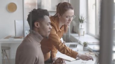 Laptop ekranında Afro-Amerikalı erkek meslektaşlarının yaratıcı projelerini inceleyen deneyimli bir beyaz uzmanın orta kavisli görüntüsü ve o dikkatle izlerken tasarım fikrini önermesi.