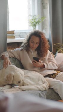 Akıllı telefon kullanan ve evdeki kanepede birlikte dinlenirken Golden Retriever köpeğini okşayan genç bir kadının dikey görüntüsü.