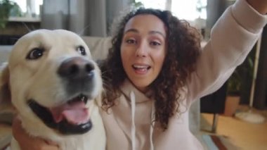 Genç, neşeli bir kadın uzanmış koluyla akıllı telefonu tutuyor ve sevimli Golden Retriever köpeğiyle fotoğraf çekerken kameraya poz veriyor.