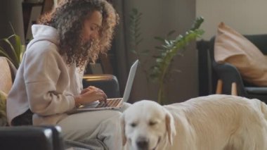 Evdeki kanepede oturan, dizüstü bilgisayarda yazan ve sonra ip oyuncağını alıp köpekle oynayan genç bir kadın.