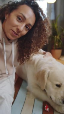 Pozitif genç bir kadının kameraya baktığı, görüntülü konuşma yaptığı ve Golden Retriever köpeğini ağzıyla kağıt şapkasını yırtarken okşadığı dikey bir görüntü.