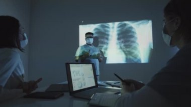 Yüz maskeli iki kadın doktor karanlık laboratuvarda dizüstü bilgisayarlarıyla oturup erkek meslektaşlarıyla Cvid-19 tedavisi üzerinde çalışırken göğüs röntgenini tartışıyorlar.