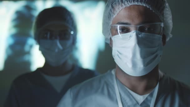 制服を着た中東の男性医師 保護メガネ 医療用マスクと帽子胸のX線投影と暗い研究室で女性同僚と立ってカメラを見て — ストック動画