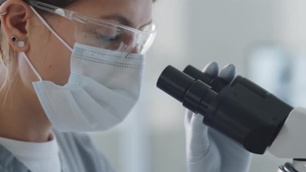 コロナウイルスパンデミック時の研究を行いながら 顕微鏡を通して見て保護面マスク 滅菌手袋で女性研究室の労働者のショットを閉じます — ストック動画