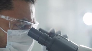Laboratuvar araştırmaları sırasında mikroskop kullanarak koruyucu yüz maskesi, gözlük ve eldiven takan orta doğulu erkek bilim adamının yakın çekim görüntüleri.