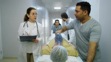 Bir grup çok ırklı doktor acil servis hastalarına valf maskesi takıyor ve hastane koridorunda sedye itiyor.