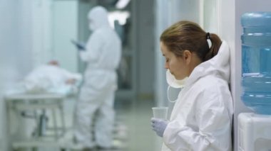 Koruyucu kıyafetli kadın doktor hastane koridorunda duruyor, duvara yaslanıyor, su içiyor ve Coronavirus salgınında fazla mesai yaparken etrafa bakıyor.