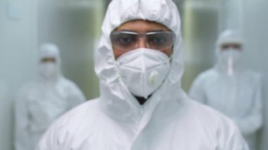 Koruyucu üniformalı, yüz kalkanı ve maskesiyle hastane koridorunda dikilip salgın sırasında kameraya bakarken kendine güvenen erkek doktorun portre fotoğrafını yakınlaştır.
