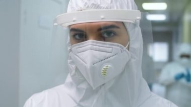 Koruyucu üniformalı, yüz kalkanı ve maskeli genç bir kadın doktorun klinikte dikilip kameraya poz verirken görüntülerini yakın çekim yapın.