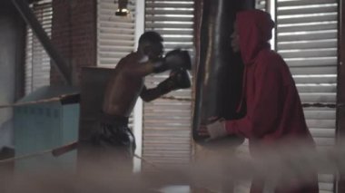 Knee-up shot of young Afro-American boxer with bare chest, in shorts and gloves vigorously punching heavy cylindrical bag at amateur boxing club and coach in hoodie holding it up