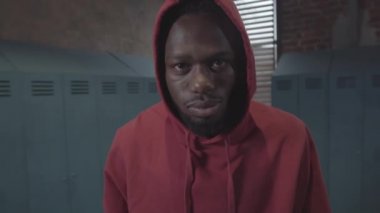Chest-up portrait shot of young African American man in hooded sweatshirt standing in dingy brick locker room of gym and staring at camera with menacing look on face