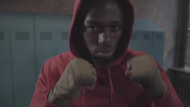 身着红色帽衫的年轻非洲裔美国拳击手的脸和手的特写照片 手套在紧握着的拳头上 站在更衣室的警戒位置上 看着镜头 — 图库视频影像