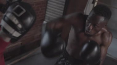 Medium high angle shot of young African male boxer with bare torso and gloves striking punch mitts upwards with forceful jabs and hooks, and black coach in hoodie holding them and giving directions