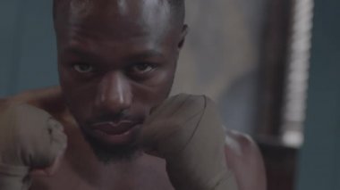 Close-up portrait arc shot of face and hands of young Afro-American MMA fighter with bare torso and boxing straps on wrists posing in locker room with clenched fists and looking at camera