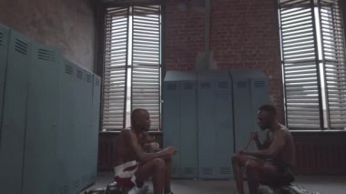 Full tilting shot of two Afro-American boxers with bare torsos, in shorts sitting in locker room of gym, wrapping wrists with hand wraps in preparation for sparring, and discussing fighting strategy