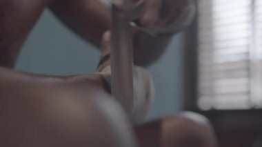 Tilting handheld close-up shot of hands and face of young black MMA fighter with bare torso and shaved head sitting in locker room and wrapping wrists in boxing hand wraps
