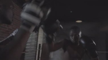 Medium handheld shot of professional young African boxer in gloves and coach with punch mitts training in boxing ring, striking with powerful jabs and hooks, ducking, and trainer laughing