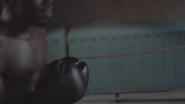 Waist-up handheld shot of young African American boxer in shorts, with bare torso and gloves furiously hitting heavy punch bag with forceful blows in rundown underground fight club