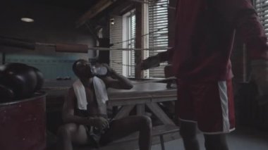 Medium shot of young African American boxer with bare torso sitting on bench next to boxing ring, sparring partner walking in, putting down gloves, both men bumping fists, drinking water and chatting