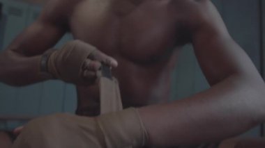 Tilting close-up shot of face, hands and bare chest of muscular young black MMA fighter wrapping wrists with boxing straps, then clenching fists, flexing arms and practicing some punching moves