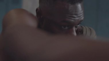 Close-up shot of young African American boxer with bare chest and hand wraps on wrists shadow boxing alone in changing room, practicing hooks, jabs and uppercuts then looking at camera