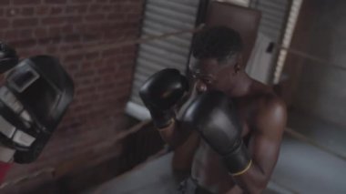 Medium high-angle shot of focused Afro-American boxer with bare chest and gloves practicing jabs on punch mitts while training with coach in dimly lit boxing gym