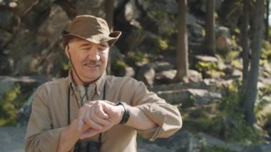 Waist up arc shot of senior man using navigation app on smartwatch during hike in nature on summer day