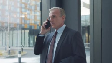 Orta yaşlı, beyaz takım elbiseli ve kravatlı bir işadamının girişteki koridorda dikilirken, sokağa bakarken ve cep telefonuyla konuşurken belden yukarı pozu.