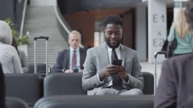 Orta boy Afro-Amerikan bir adamın iş elbisesi ve kravatıyla havaalanının kalkış salonundaki koltukta otururken, başka tarafa bakarken, gülümserken ve akıllı telefondan yazarken ve bekleyen diğer yolcular.