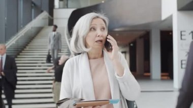 Göğsü kabaran orta yaşlı, gri saçlı, havaalanında yürüyen, biniş kartı tutan, cep telefonuyla sohbet eden ve arka planda sarılan bavulu olan neşeli, orta yaşlı, melez bir kadının görüntüsü.