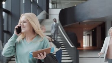 Orta yaşlı, sarışın bir kadının uçuş kartı ve pasaportuyla havaalanında yürürken, cep telefonuyla konuşurken, gülümserken ve bavul taşıyan yolcuların göğüs üstü çekimleri.