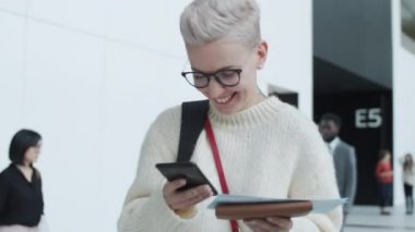 Göğüs hizasında, kısa sarı saçlı, şık genç beyaz bir kadının kalabalığın arasında dikilirken, elinde akıllı telefon ve uçuş bileti tutarken, mesaj yazarken, gülerken ve etrafa bakarken.