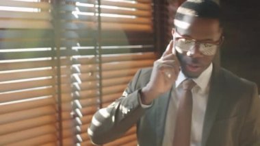 Resmi takım elbiseli ve gözlüklü Afro-Amerikan işadamının göğsüne bakın. Pencerenin yanında duruyor. Ofisinde panjurlar var ve cep telefonuyla konuşuyor.