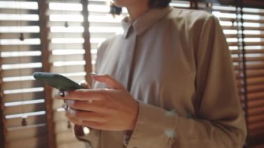 Şık bluz giyen bir iş kadınının pencerenin yanında güneş ışığıyla dolu panjurlarla dururken akıllı telefonuyla mesajlaştığı orta bölüm görüntüsü.