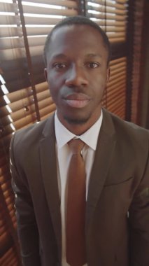 Dikey bel üstü çekimde, resmi takım elbiseli yakışıklı Afro-Amerikan iş adamı, pencerenin yanında duruyor, gülümsüyor ve kameraya bakıyor.