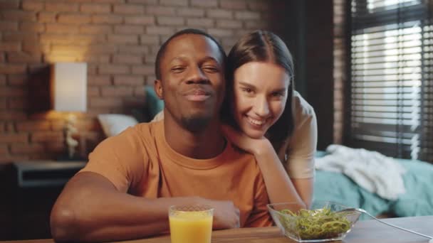 年轻的黑人男人坐在桌旁 在家里拿着沙拉和果汁 给他的白人妻子打电话 快乐的夫妻一边挥手一边笑一边在摄像机前聊天 — 图库视频影像