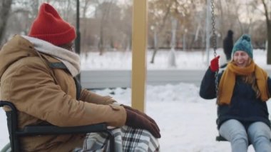 Kış yürüyüşü sırasında parkta salıncakta sallanırken tekerlekli sandalyede gülümseyen ve bayan arkadaşıyla konuşan neşeli Afro-Amerikalı adamın arkası.