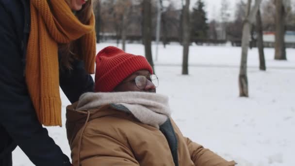 車椅子でアフリカ系アメリカ人男性の友人を押して 冬の日に屋外に歩いている間に彼と何かを話し合う若い白人女性 — ストック動画