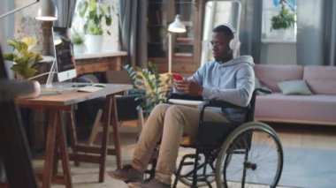 Tekerlekli sandalyedeki belden aşağısı felçli Afro-Amerikalı genç adam akıllı telefon kullanıyor ve evde kablosuz kulaklıkla müzik dinliyor.