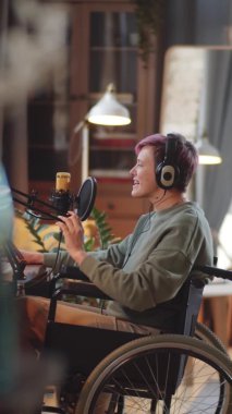 Tekerlekli sandalyedeki, kablosuz kulaklıklı, mikrofona konuşan belden aşağısı felçli genç bir kadının ev stüdyosunda podcast yaparken çekilmiş dikey görüntüleri.