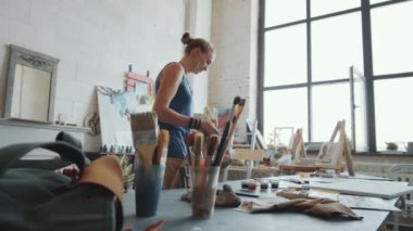 Genç bayan sanatçı sırt çantasını masaya bırakıyor, önlüğünü giyiyor ve stüdyoda sehpaya gidiyor.