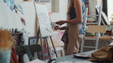 Profesyonel kadın sanatçının stüdyoda sehpanın başında durup tuvale soyut bir resim çizmesinin orta bölümü.