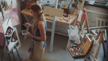 Önlüklü, palet tutan ve resim yapan genç sarışın bir kadının sanat stüdyosunda çalışırken resmini yüksek açılı çekimleri.