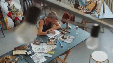 Profesyonel bir kadın sanatçının stüdyoda masa başında oturup pastel boyalarla resim yaptığı yüksek açılı bir fotoğraf.