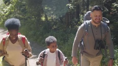 Neşeli Afro-Amerikan anne, beyaz baba ve küçük melez oğul güneşli yaz gününde ulusal parkta yürüyüş direkleri ve sırt çantalarıyla birlikte yürürken gülümsüyor ve konuşuyorlardı.