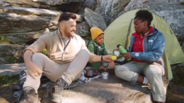 Neşeli Afro-Amerikalı kadın yaz günü kamp alanında dinlenirken beyaz koca ve küçük melez oğlu için termostan fincanlara çay dolduruyor.