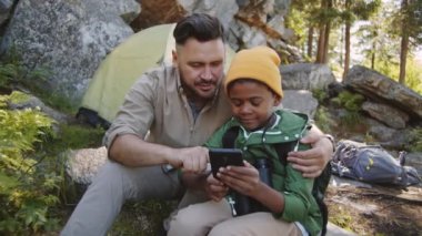 Neşeli Kafkasyalı bir adam, küçük melez oğlunu kucaklıyor, akıllı telefon ekranına bakıyor, sohbet ediyor ve çocukla birlikte kamp alanında otururken gülümsüyor.