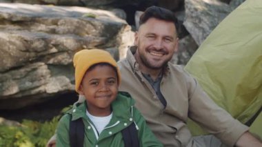 Neşeli beyaz adamın portresi ve onun küçük melez oğlu yaz günü kamp alanında birlikte oturur, kameraya bakar ve mutlu bir şekilde gülümserler.