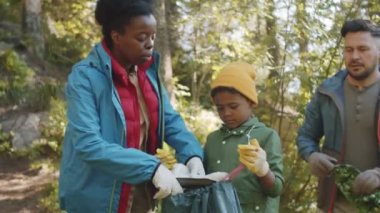 Küçük melez ırk çocuğu çöp torbası tutarken ailesi ormanda kamp yaptıktan sonra içine plastik atık koyuyor.